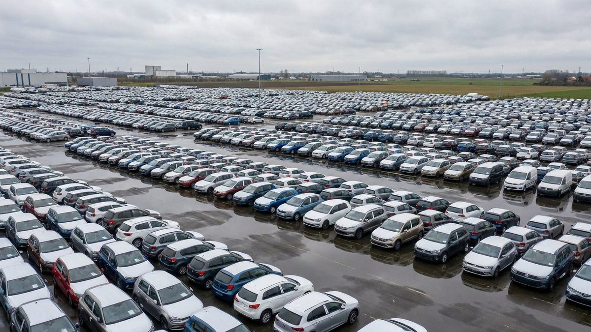 Vaste parc de stockage de voitures neuves de plusieurs marques alignées sous un ciel gris